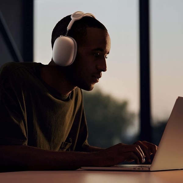 A high-contrast, moody close-up of a person wearing minimalist over-ear headphones while working on a laptop in a dimly lit environment.