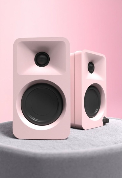 A high-quality, studio-style product shot of two pastel pink Bluetooth speakers placed on a soft grey circular pedestal against a solid pink background.