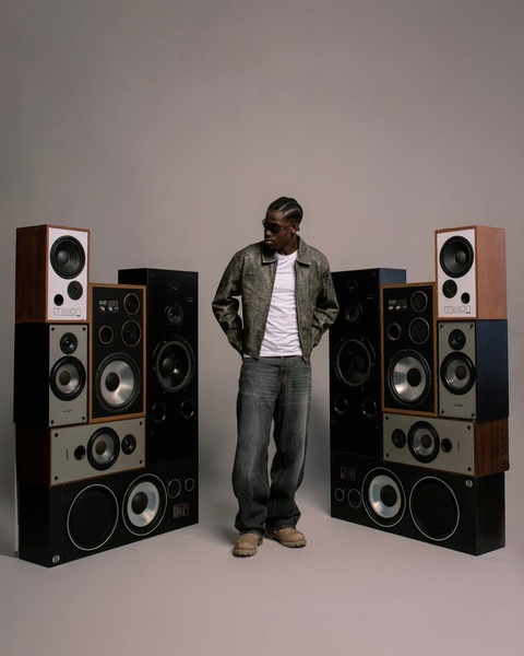 A stylish, centered shot of a model standing between a symmetrical arrangement of stacked vintage speakers against a minimalist studio background.