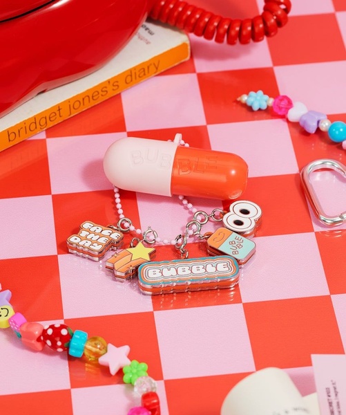 A high-contrast, playful product shot of earbuds in a capsule-shaped case, arranged with colorful charms on a checkered surface.
