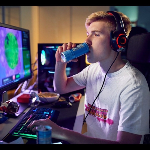 A gamer is deeply engaged in a late-night session, holding an energy drink while wearing gaming headphones in a dark room with neon accents.
