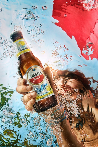 A high-contrast, underwater-themed product shot of an Amstel Radler bottle featuring dynamic water bubbles and vibrant red accents.