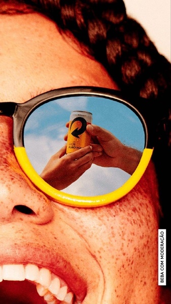A close-up shot of a person wearing yellow-rimmed sunglasses, with a summer sky and a canned drink being toasted visible in the reflection.