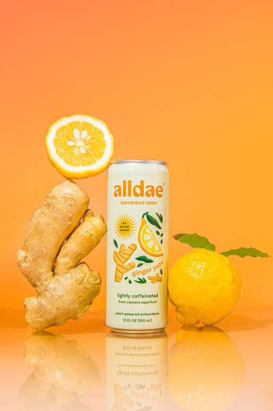 A studio-quality product image of a Superfruit Soda can balanced with fresh ginger and yuzu, set against a vibrant solid orange background.