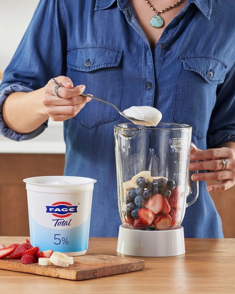 A close-up shot of a person adding Greek yogurt to a blender filled with fresh fruit, emphasizing the natural ingredients of the product.