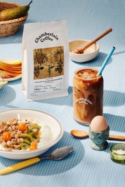 A high-quality, top-down view of a balanced morning meal featuring a cold brew coffee with a branded glass, granola bowl, and a soft egg, styled on a blue grid tablecloth.