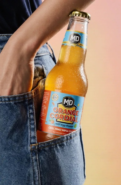A close-up lifestyle shot of a glass bottle of orange cordial tucked into a person's jeans pocket, highlighting the product as an everyday essential.
