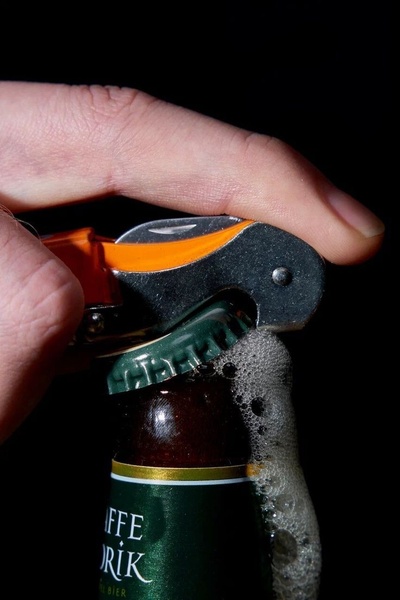 A sharp, macro-style image focusing on a hand using an orange opener to pry the cap off a dark green beer bottle, capturing the moment of carbonation and foam.