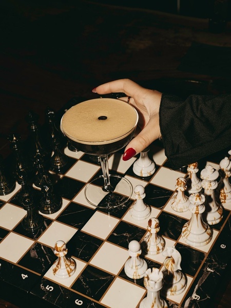 A high-contrast, moody close-up of a hand holding an espresso martini glass over a marble chessboard, emphasizing a luxury lifestyle.