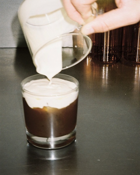 A close-up aesthetic shot of cream being poured into an iced coffee, focusing on the visual layering and texture of the drink.