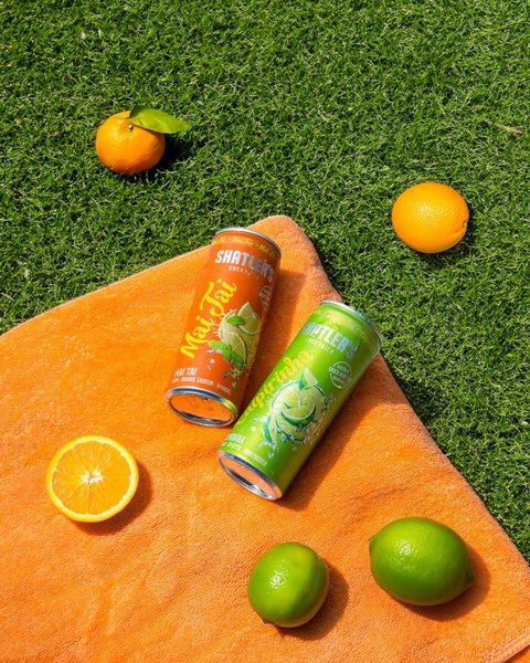 Two cans of cocktail drinks are displayed on an orange towel placed on a lush green lawn, surrounded by fresh whole and sliced oranges and limes.