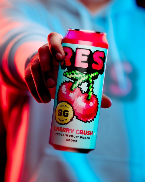 A close-up shot of a person holding a pixel-art style Cherry Crush protein drink can against a high-contrast red and blue neon light background.