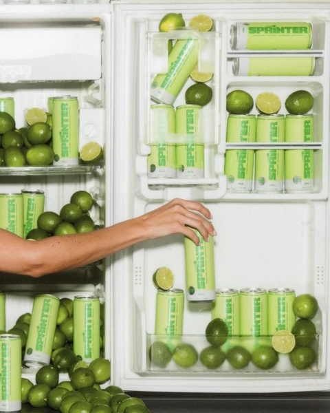 A high-contrast photograph of a refrigerator fully stocked with lime soda cans, with a hand reaching in to grab one, emphasizing product availability and refreshment.
