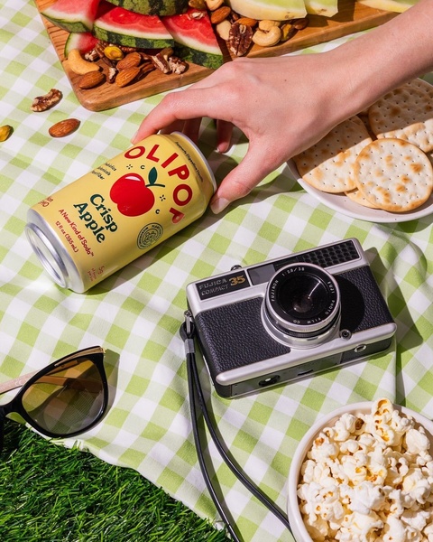 A high-angle, lifestyle-focused shot of a Crisp Apple soda can placed on a picnic blanket surrounded by snacks and a vintage camera.