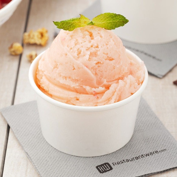A close-up studio shot of a single scoop of fruit-flavored ice cream in a simple paper cup, garnished with a fresh mint leaf.
