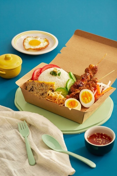 A high-quality, top-down-angled studio photograph of a takeaway meal box featuring rice, grilled skewers, sliced eggs, and fresh vegetables on a vivid blue background.