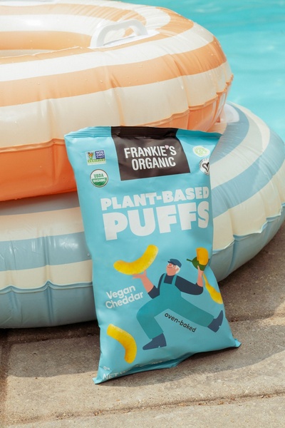A bright, outdoor-themed product shot featuring a bag of Plant-Based Puffs resting against inflatable pool floats.
