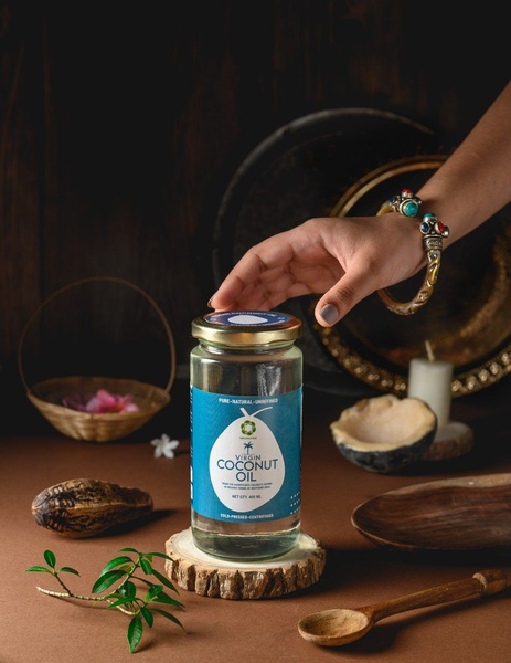 A high-end, moody product shot of a Virgin Coconut Oil jar staged with natural elements like wood, leaves, and a coconut shell, captured from a 3:4 aspect ratio perspective.