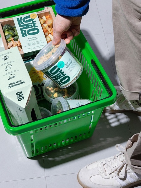 A top-down, first-person view of a person placing ready-to-eat salad containers into a vibrant green grocery basket to demonstrate product convenience.