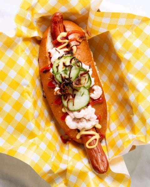 A high-quality, top-down shot of a gourmet hot dog served in a yellow gingham paper basket, focusing on fresh ingredients and vibrant textures.