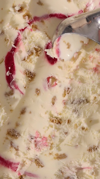 A close-up shot capturing a metal scoop pulling through creamy ice cream, revealing ribbons of berry swirl and cookie crumble inclusions.