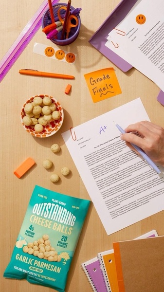 A top-down, flat-lay photography style showing a bag of cheese balls alongside study materials like a graded paper and school supplies.