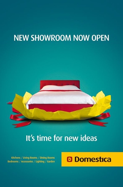 A minimalist studio shot of a bed gift-wrapped on a solid teal background, announcing a new furniture showroom opening.