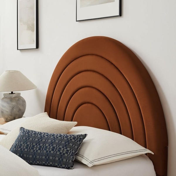 A close-up, high-aesthetic shot of a rust-colored velvet headboard with a geometric arch design, styled with neutral bedding and a textured lamp.