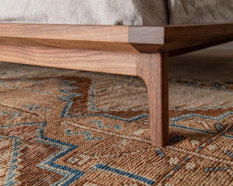 A close-up, macro-style product shot of a walnut bed frame focusing on the joinery, texture, and natural wood grain against a woven rug.