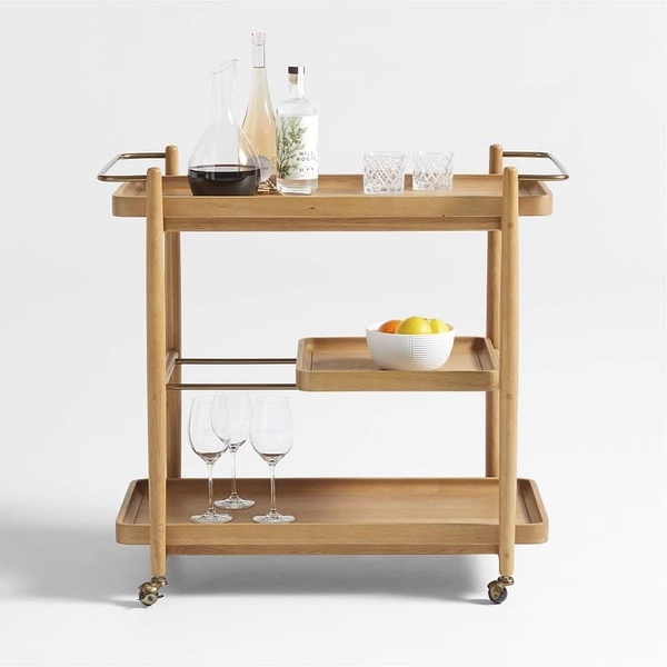 A clean, high-end studio product shot of a wooden bar cart displaying glassware and bottles. The setup focuses on lifestyle aspiration through minimalist staging.