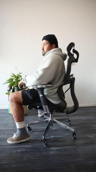 A side-profile shot of a person sitting in an ergonomic office chair, focusing on the chair's lumbar support and overall design in a natural home environment.