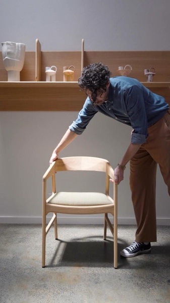 A clean, studio-lit photograph featuring a designer wooden chair being carefully adjusted by a professional in a warm, neutral environment.
