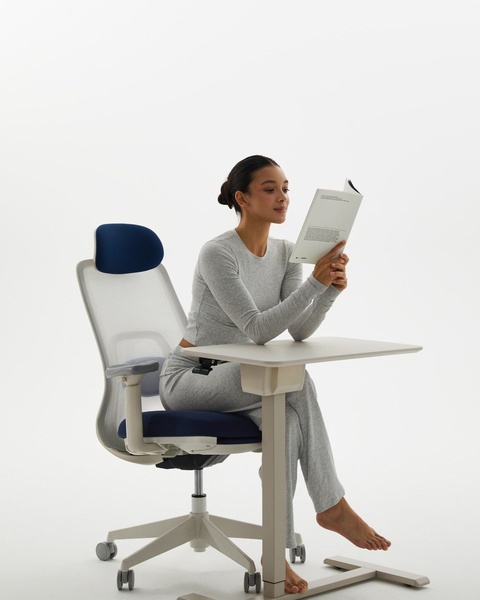 This creative features a woman relaxing in a modern, ergonomic desk chair while reading, emphasizing a lifestyle-focused, high-comfort positioning.