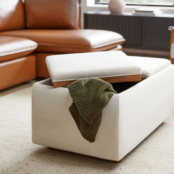 A high-quality lifestyle photograph of a minimalist cream-colored storage ottoman featuring a wood-topped lid with an olive green throw blanket.