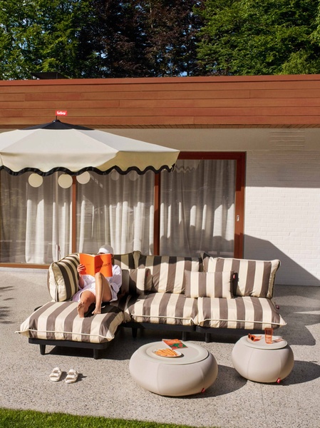 A high-end lifestyle shot of a woman reading on a striped modular outdoor sofa set under a matching scalloped patio umbrella in a sun-drenched courtyard.