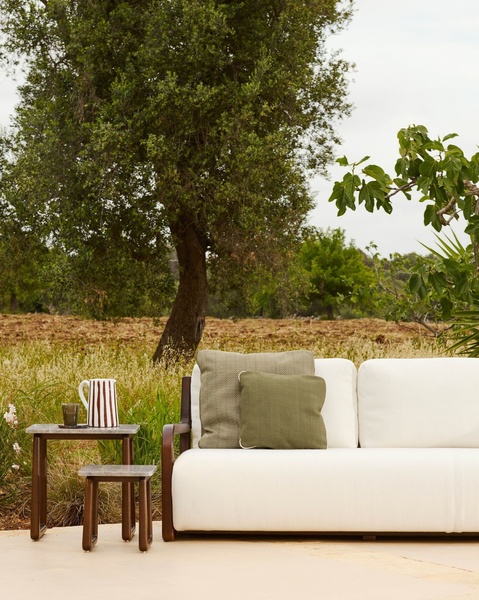 A high-end outdoor sofa paired with textured cushions and a small accent table, set against a natural rustic landscape.