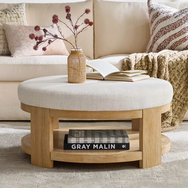 A clean, lifestyle-focused shot of a neutral-toned round wooden coffee table styled in a sunlit living room setting.