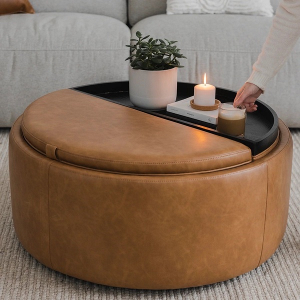 A high-quality lifestyle shot of a round leather storage ottoman styled in a neutral home interior, featuring a candle and plant to evoke warmth.