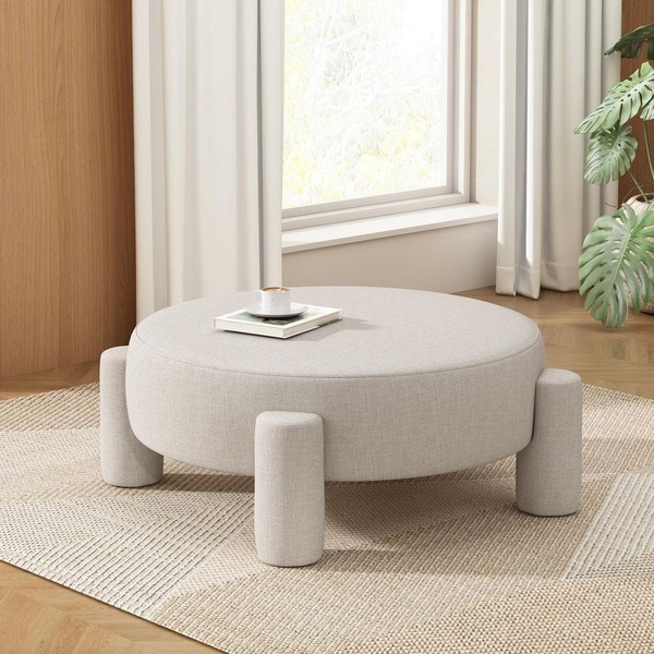 A high-quality, clean-background shot of a modern, low-profile round ottoman-style coffee table styled in a sunlit room.