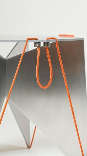 This creative showcases a sleek metal stool with orange cord detailing, focusing on texture and material contrast in a clean, high-end studio setting.
