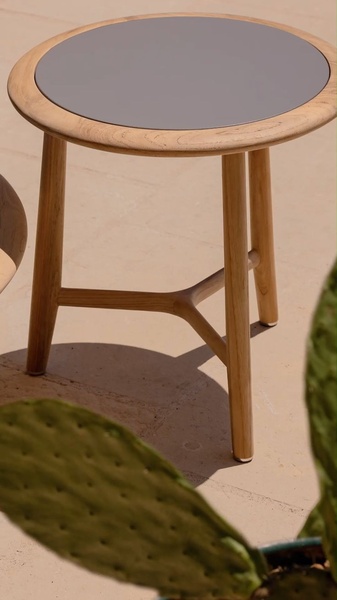 A high-quality, sun-drenched close-up shot of a round wooden side table placed on a patio, highlighting its texture and material quality.