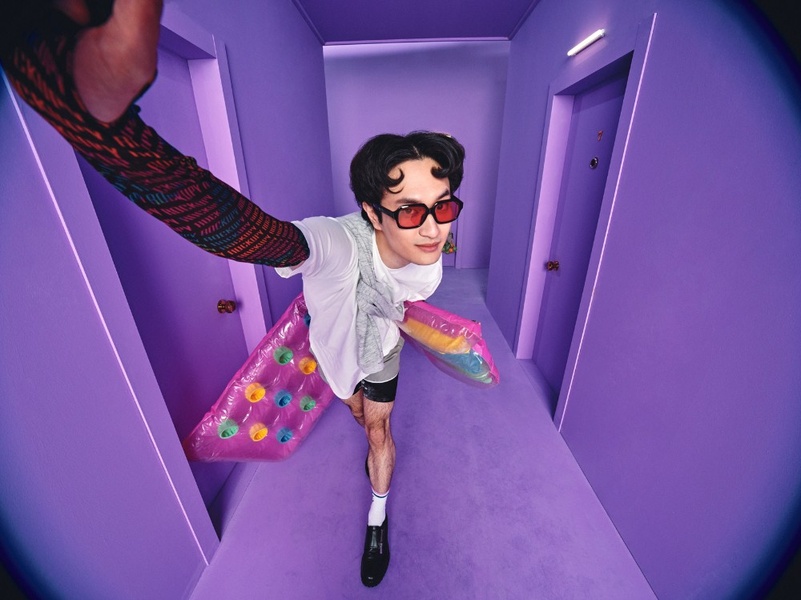 A high-angle, wide-lens shot of a man wearing red-tinted glasses and a shirt with branded sleeves, holding a bright pink inflatable pool float inside a solid violet-colored corridor.