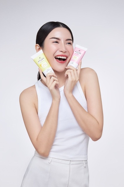A high-energy, clean beauty ad featuring a model holding two skincare tubes against a neutral studio backdrop.