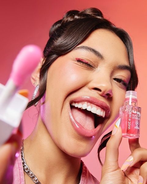 A close-up portrait of a model winking while holding a vibrant lip oil, utilizing a high-energy, friendly aesthetic.