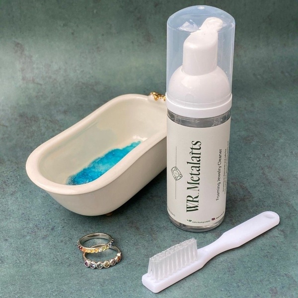 A high-angle 1:1 shot featuring foaming jewelry cleaner, a tiny bathtub prop, and a cleaning brush to visualize the product application process.