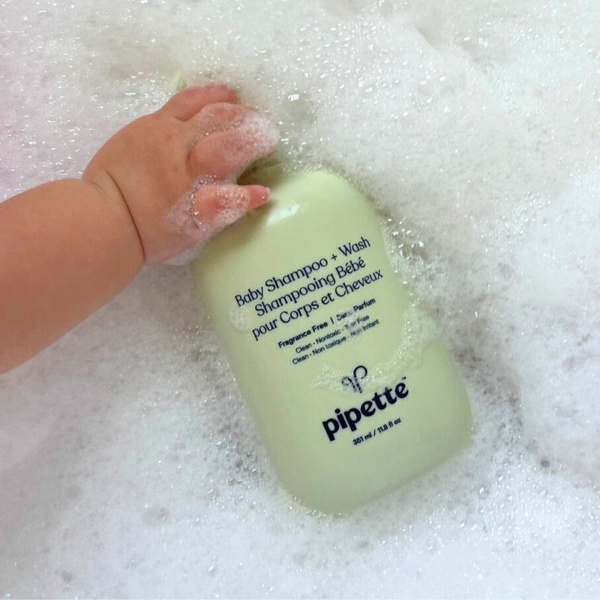 A close-up, high-angle shot of a baby shampoo bottle resting in bubbly bath water, held by a small hand to emphasize gentleness.