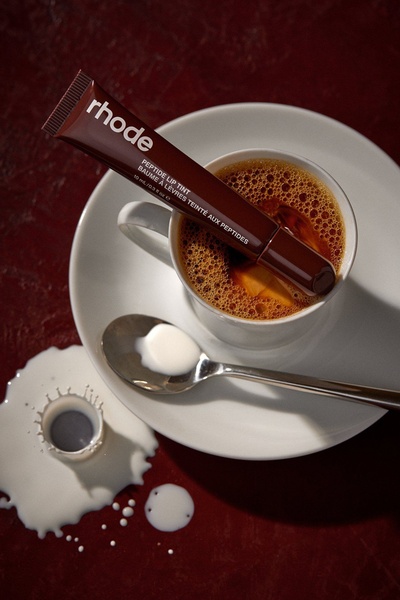 A sophisticated, high-contrast lifestyle shot featuring a peptide lip tint tube resting inside a filled espresso cup, surrounded by spilled milk and coffee.