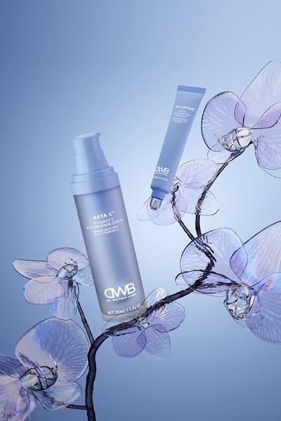 A high-end studio shot featuring a Vitamin C serum bottle and a lip treatment tube floating amidst transparent, crystalline flower structures against a soft blue gradient background.