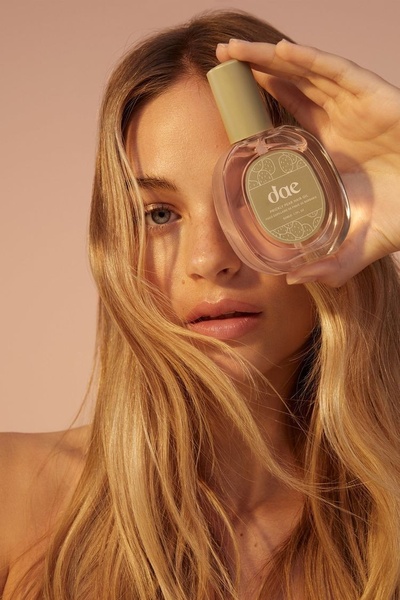 A close-up portrait of a woman holding a hair oil bottle partially obscuring her face, creating a soft, aspirational beauty aesthetic.