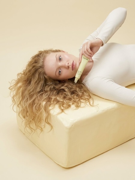 A high-end studio portrait of a model holding a lip balm while lying on a soft, butter-colored textural block to emphasize product softness.